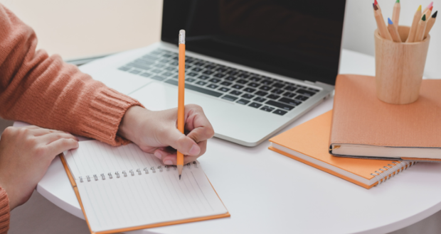 Person writing in a notebook with a computer in the background